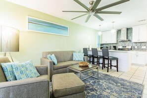 Un salon avec un canapé, un pouf et une table basse, une cuisine avec des tabourets de bar et un ventilateur de plafond.