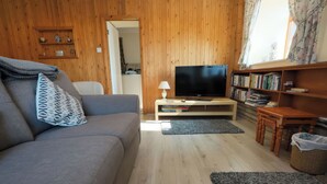 Un accogliente soggiorno con un divano grigio, una televisione a schermo piatto su un supporto in legno, una libreria piena di libri e un pavimento in legno.