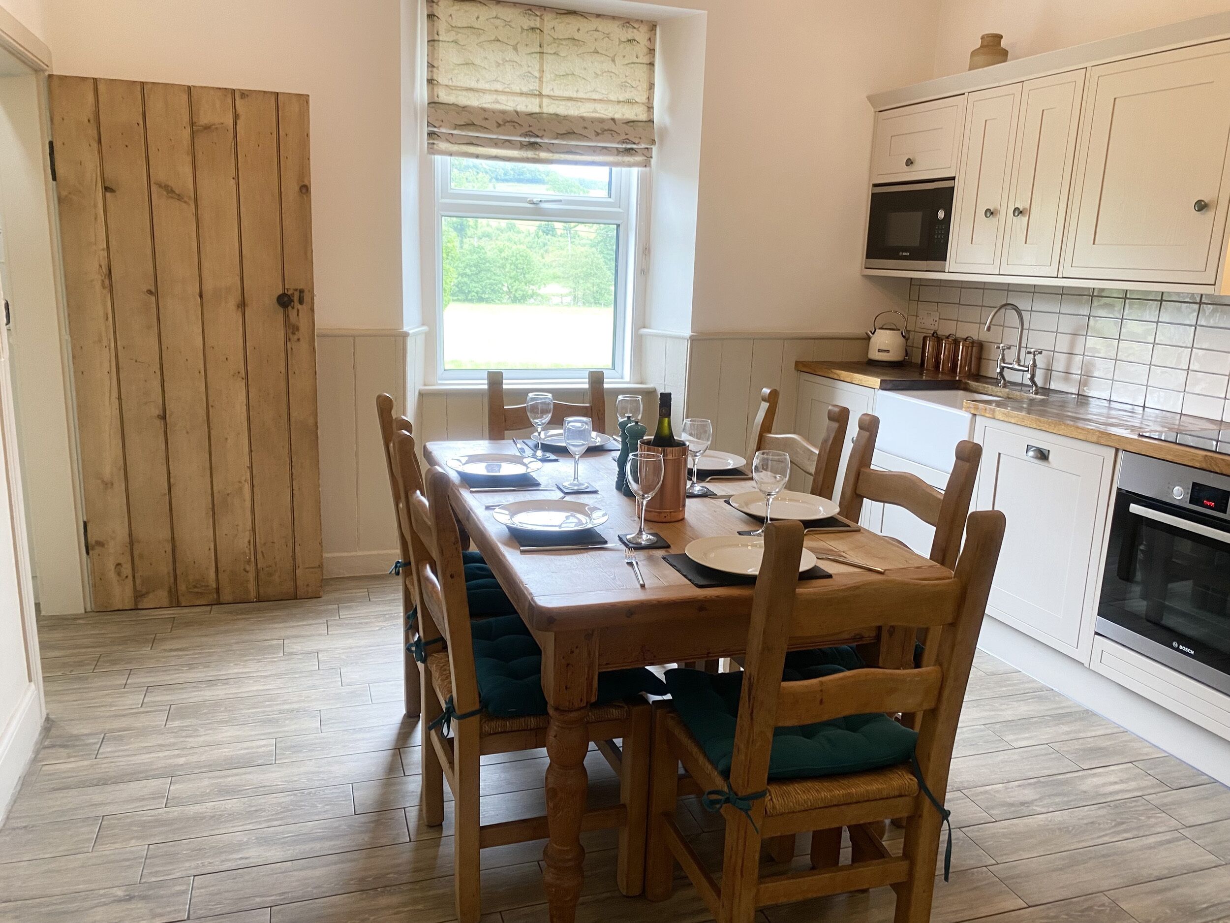 A dining area with a wooden table set for four, surrounded by wooden chairs with green cushions. A window with a patterned curtain allows natural light into the room.