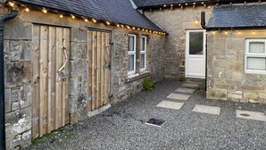 A stone building with wooden doors and windows, a gravel driveway, and string lights. - Netherraw (Hawick)
