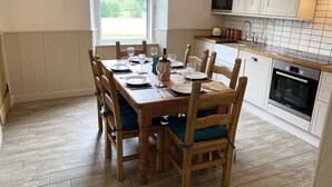A dining area with a wooden table set for four, surrounded by chairs with cushions. A window provides natural light, and a kitchen with white cabinets and a microwave is visible in the background. - Netherraw (Hawick)