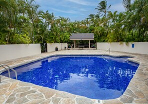 A swimming pool with a tiled deck, surrounded by a white wall and lush tropical vegetation.