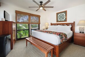 A bedroom with a bed, a wooden bench, a ceiling fan, a window with a view, and a bedside table with a lamp.