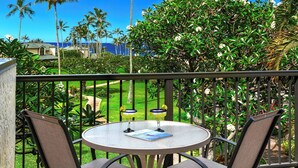 Een ronde tafel met twee stoelen, een boek en twee gele drankjes op een balkon met uitzicht op de oceaan en palmbomen.