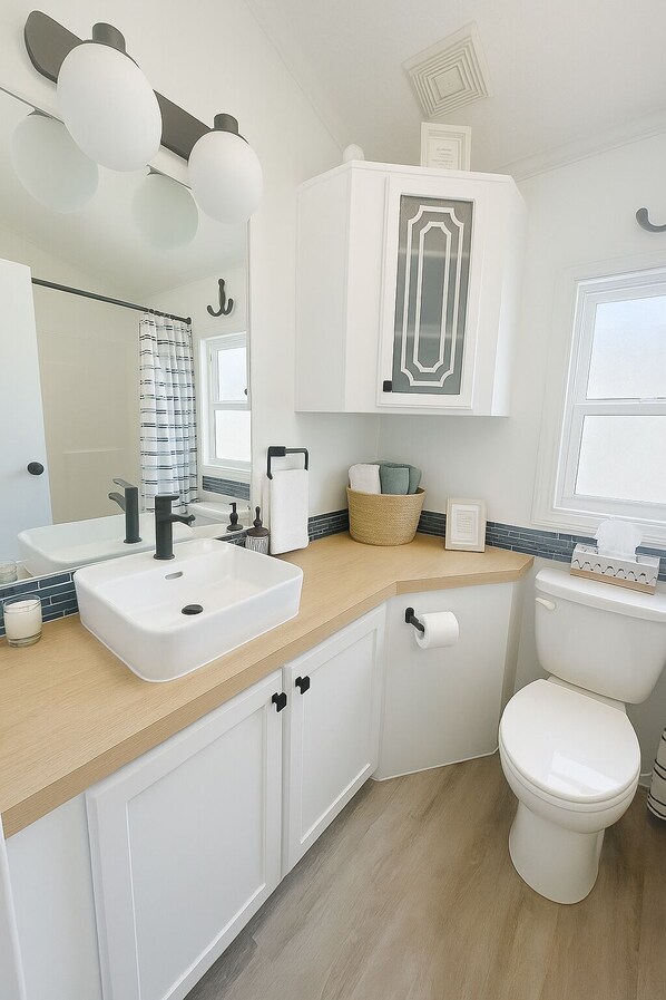 A modern bathroom with a white sink, black faucet, and a toilet. There is a mirror above the sink and a window with a view of the outside. - LAKEFRONT Park Model at Christina Lakeside Resort (Christina Lake)