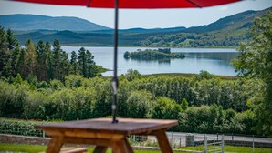 A wooden table under a red umbrella, overlooking a lake with mountains in the background.