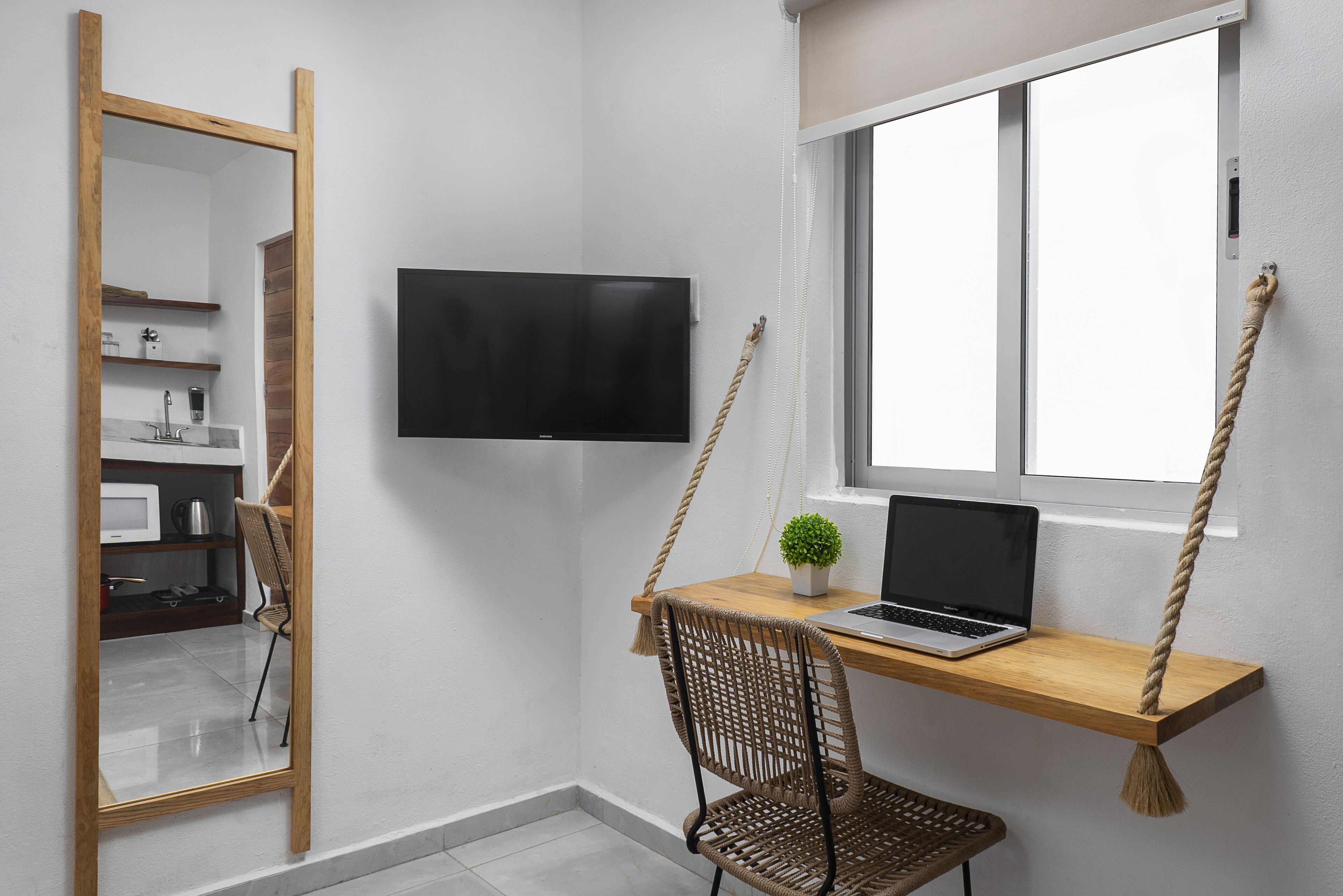 Quarto compacto com escrivaninha de madeira, um laptop, uma cadeira, TV fixada na parede e janela com balanço de corda.