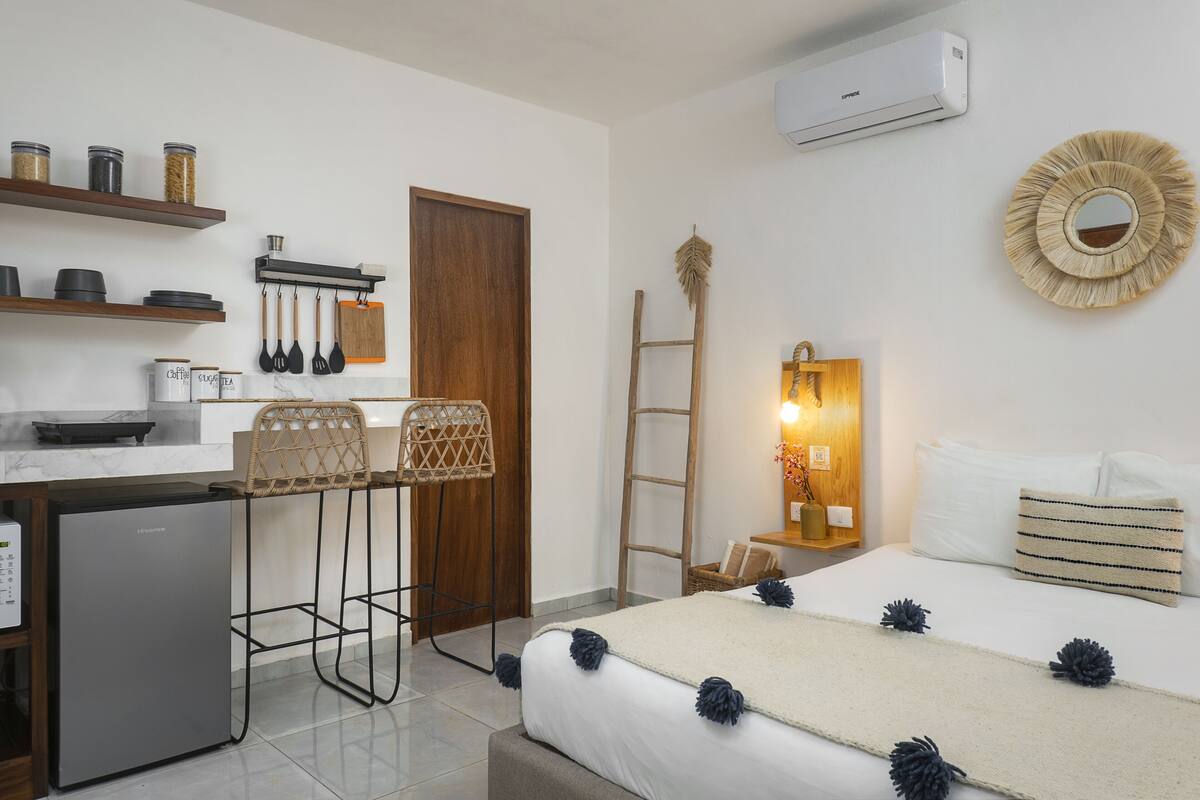 A compact hotel room with a kitchenette, a bed with decorative pillows, a small fridge, and a wooden ladder.