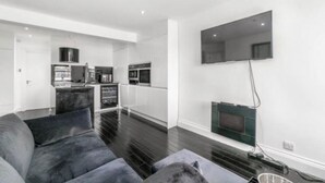 A modern living room with a grey sofa, a flat-screen TV mounted on the wall, and a kitchen in the background. - Flat close to Portobello Market (London)