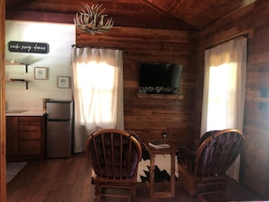A cozy cabin-style room with wooden walls, a kitchenette, a television mounted on the wall, and two chairs facing a small table.