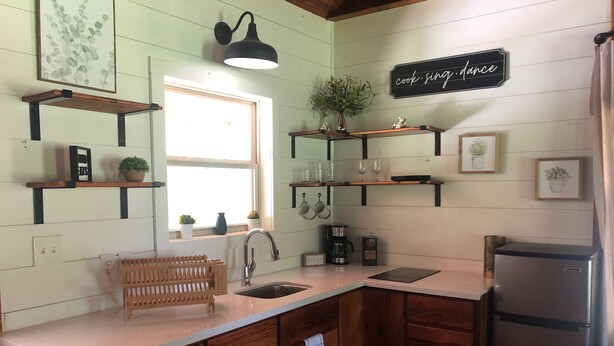 A kitchen with wooden cabinets, a stainless steel refrigerator, a sink, and a window. There are shelves with plants and kitchenware.