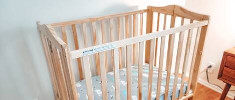 A wooden crib with a safety label, positioned in a room with wooden flooring and a white wall.
