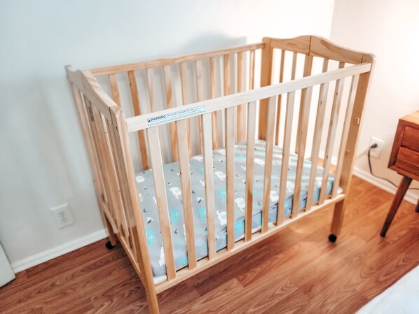 A wooden crib with a safety label, positioned in a room with wooden flooring and a white wall.