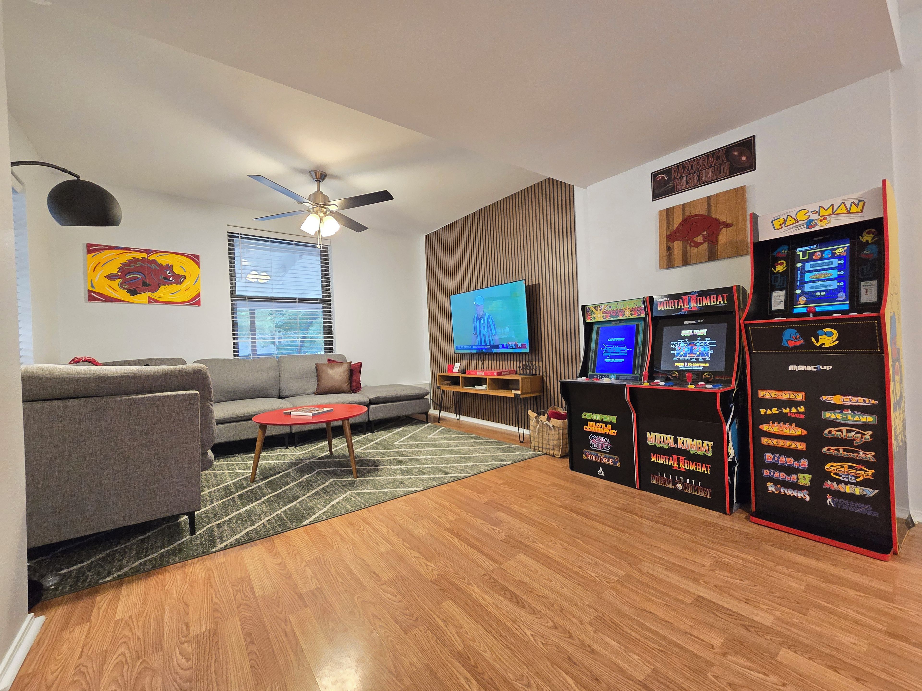Una sala de estar con un sofá gris, una mesa de centro roja, un ventilador de techo, un televisor de pantalla plana y juegos de arcade.