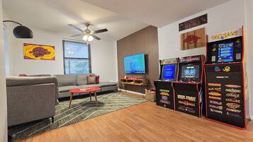 Una sala de estar con un sofá gris, una mesa de centro roja, un ventilador de techo, un televisor de pantalla plana y juegos de arcade.