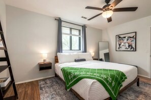 A bedroom with a bed, a ceiling fan, a window with curtains, a bedside table with a lamp, a framed picture on the wall, and a wooden floor.