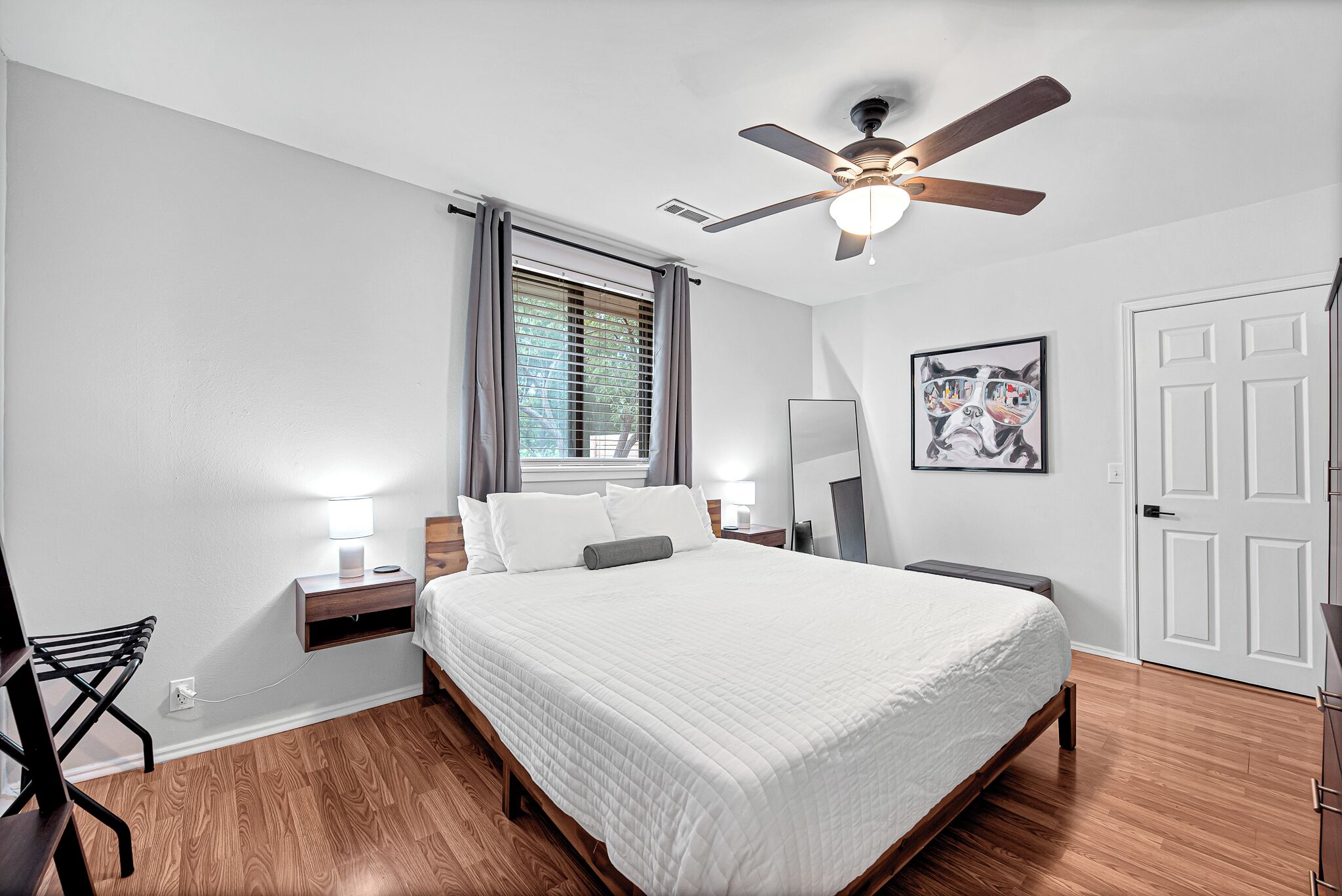 Ein Schlafzimmer mit Bett, Deckenventilator, Fenster mit Vorhängen, Nachttisch mit Lampe und einer Tür.