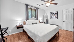 A bedroom with a bed, a ceiling fan, a window with curtains, a bedside table with a lamp, and a door.