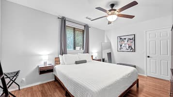 Ein Schlafzimmer mit Bett, Deckenventilator, Fenster mit Vorhängen, Nachttisch mit Lampe und einer Tür.