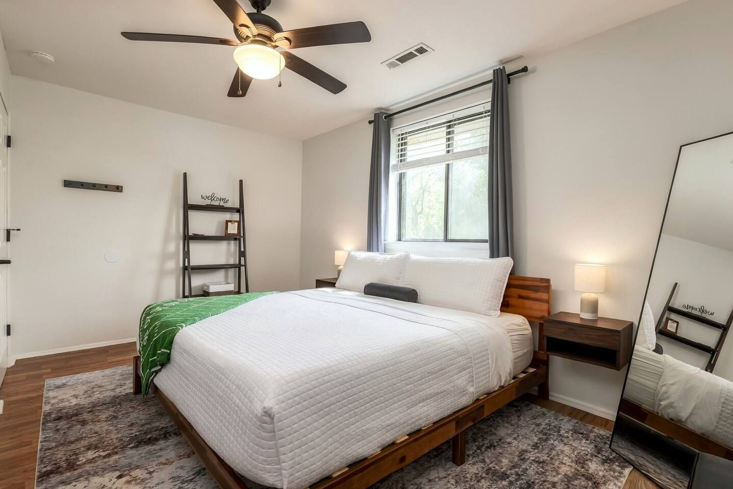 Un dormitorio con cama, ventilador de techo, ventana con cortinas, mesita de noche con lámpara y estante en la pared.