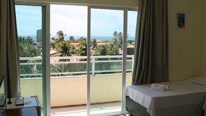 Une chambre d’hôtel comprenant un lit, un bureau avec une chaise et une grande porte-fenêtre coulissante donnant sur un balcon avec vue sur les palmiers et les immeubles.