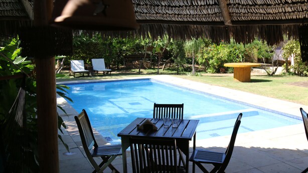 Un espace piscine agrémenté d’une table et de chaises en bois, d’un toit de chaume et offrant une vue sur la piscine.