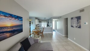 Un salon avec un canapé, un coin repas avec des chaises, une cuisine à l’arrière-plan et un tableau représentant un coucher de soleil accroché au mur.