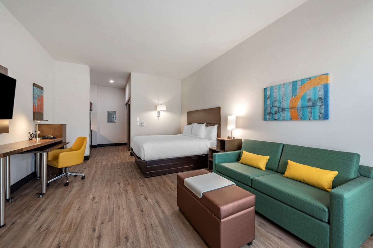 A hotel room with a bed, a green sofa with yellow pillows, a brown ottoman, a desk with a chair, and a television on the wall.