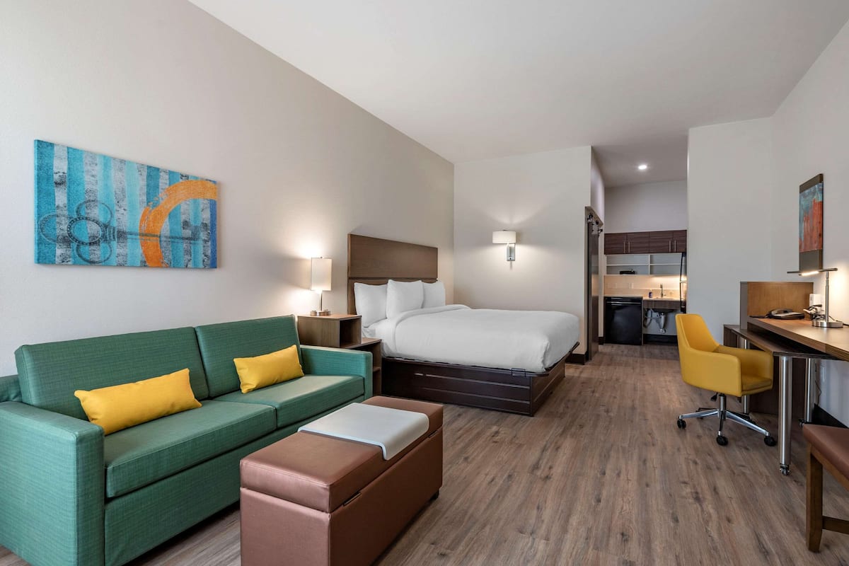 A hotel room with a green sofa, a bed, a desk, and a kitchenette in the background.