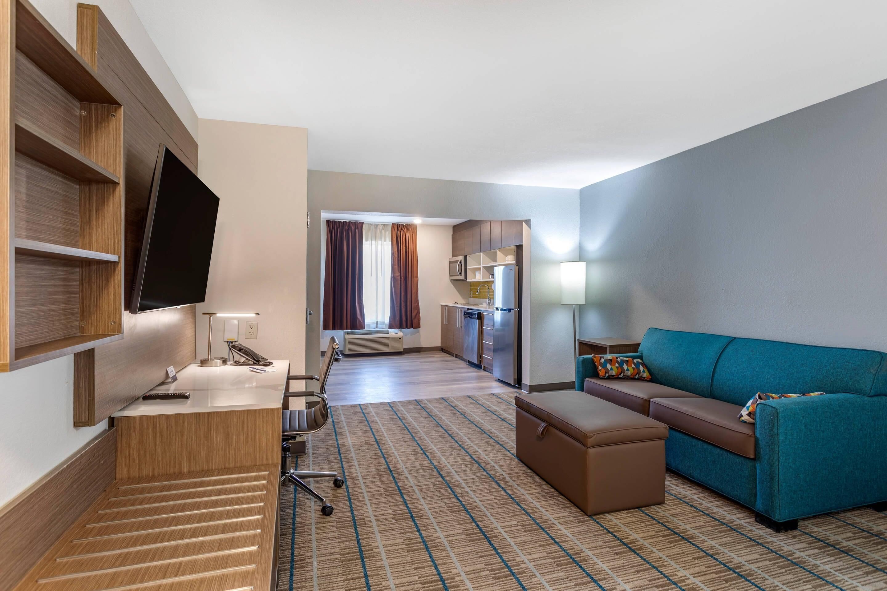 Hotelkamer met een flatscreen-tv, een bureau met lamp, een bank, een klein voetenbankje, een kitchenette en uitzicht door een raam.