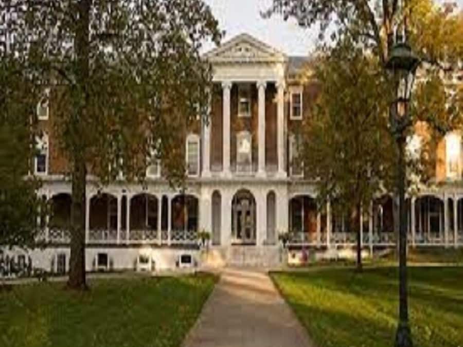 A large building with a central entrance, surrounded by trees and a pathway leading to it.