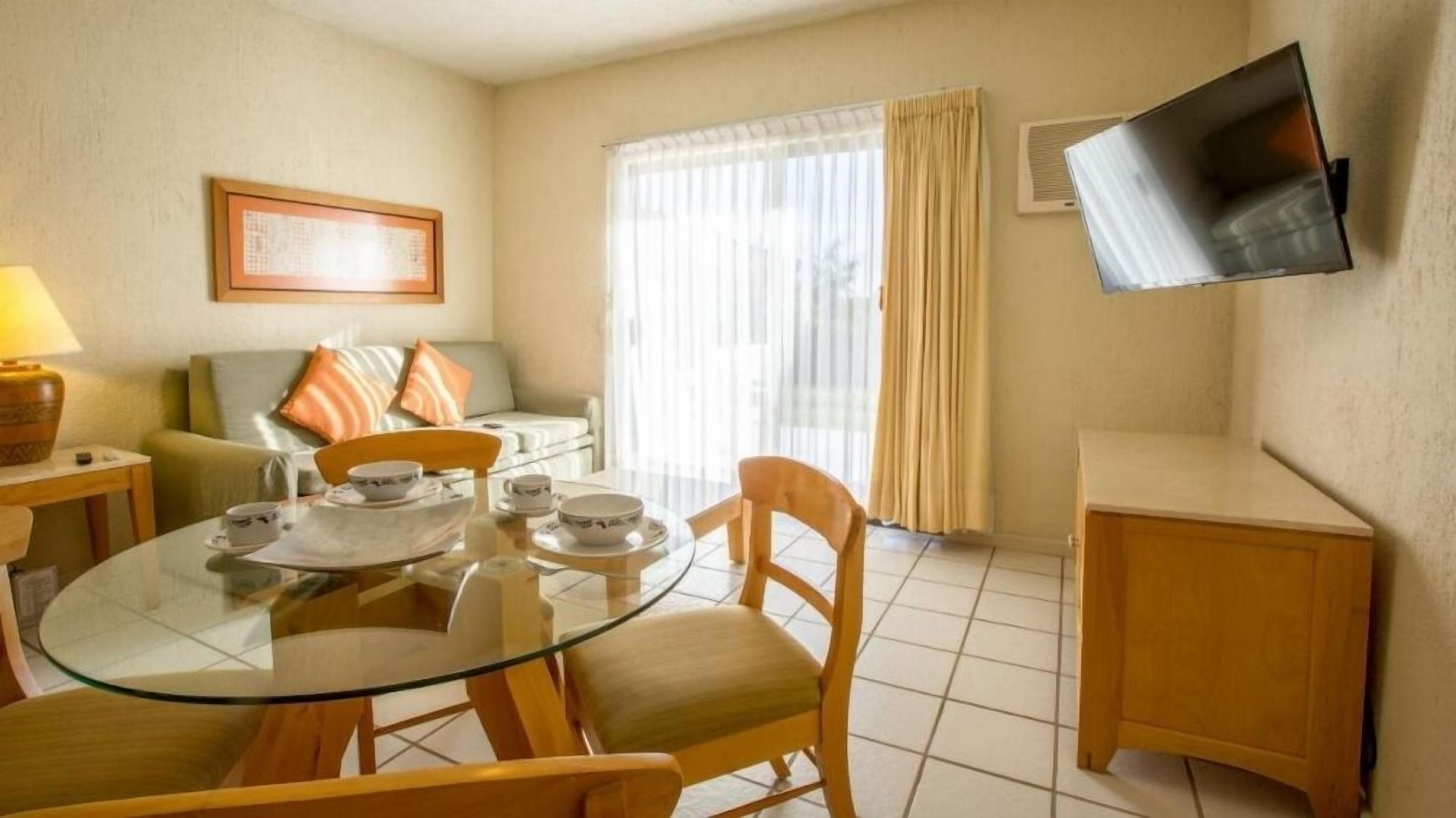 A hotel room with a glass dining table, wooden chairs, a sofa with cushions, a flat-screen TV mounted on the wall, and a window with curtains.