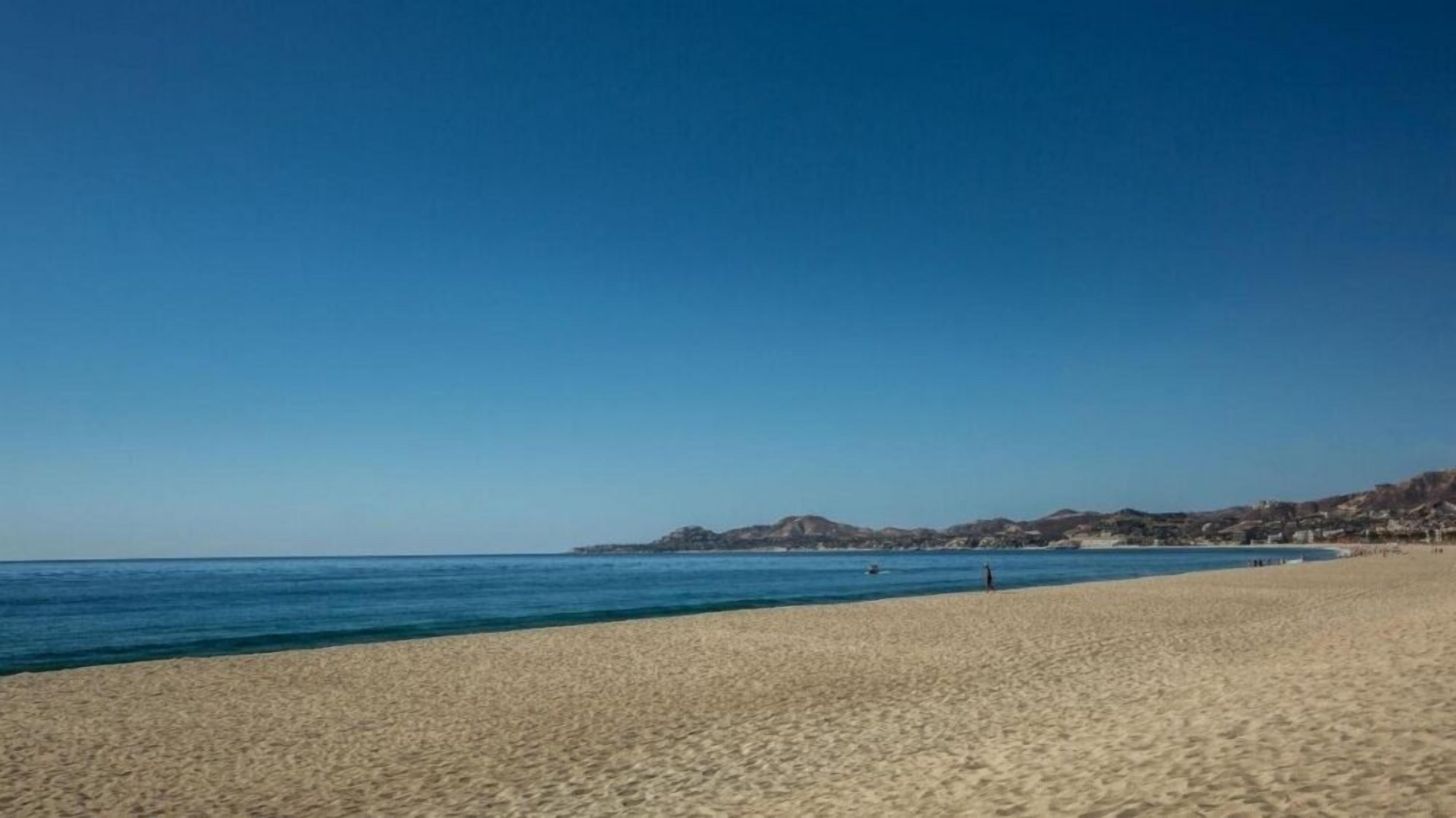 A wide sandy beach with clear blue water and distant hills.