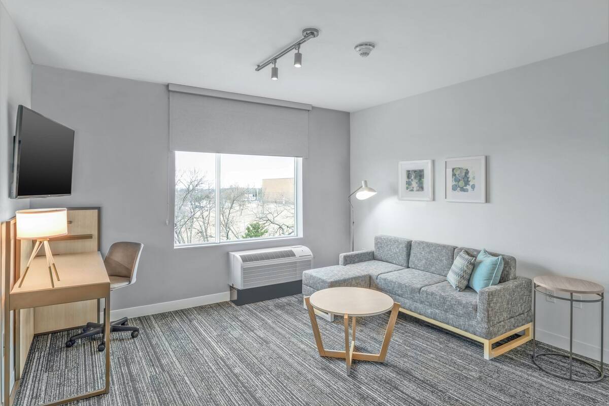 A modern living room with a grey sofa, a round coffee table, a desk with a lamp, and a TV mounted on the wall.