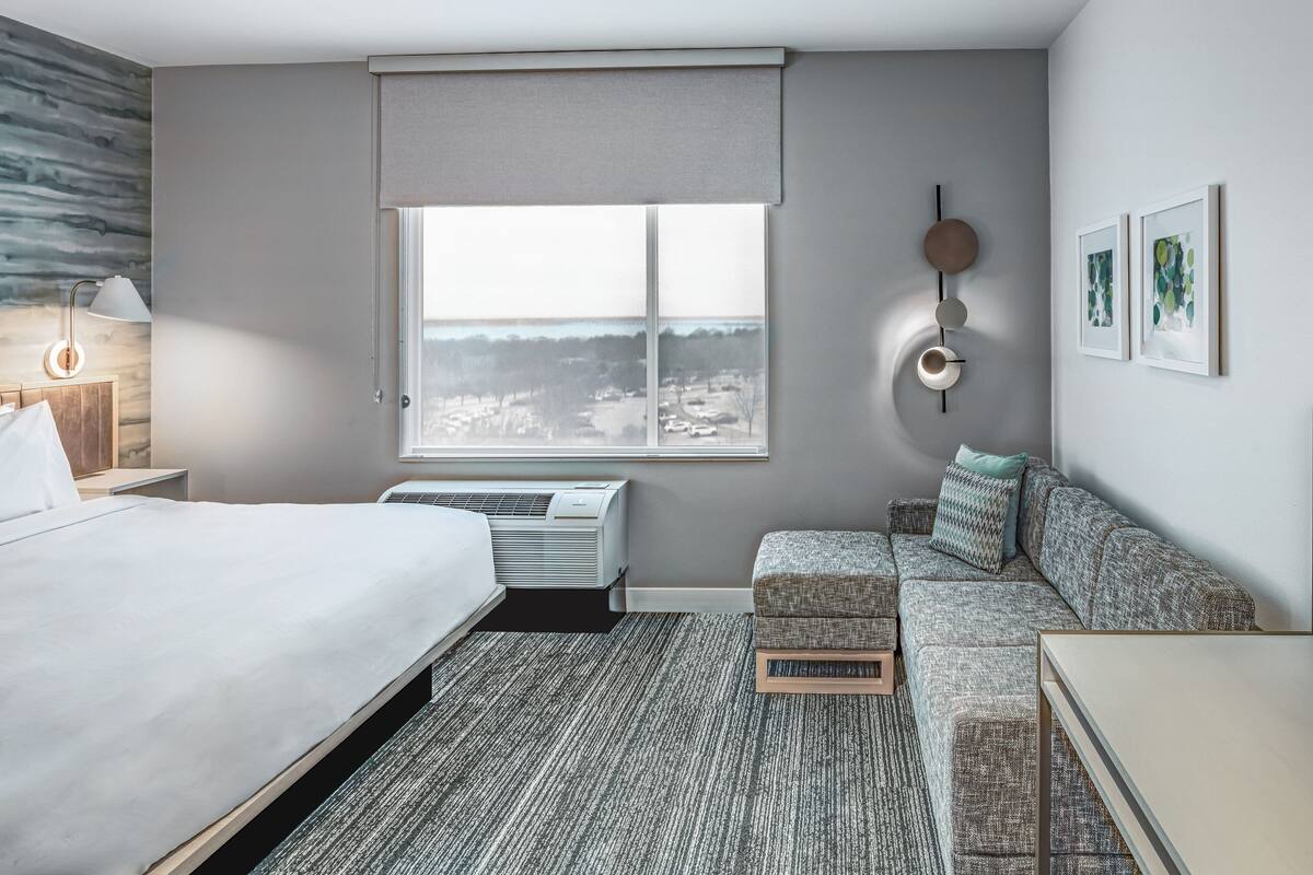 A modern hotel room with a large bed, a sofa, a small table, and a window with a city view.