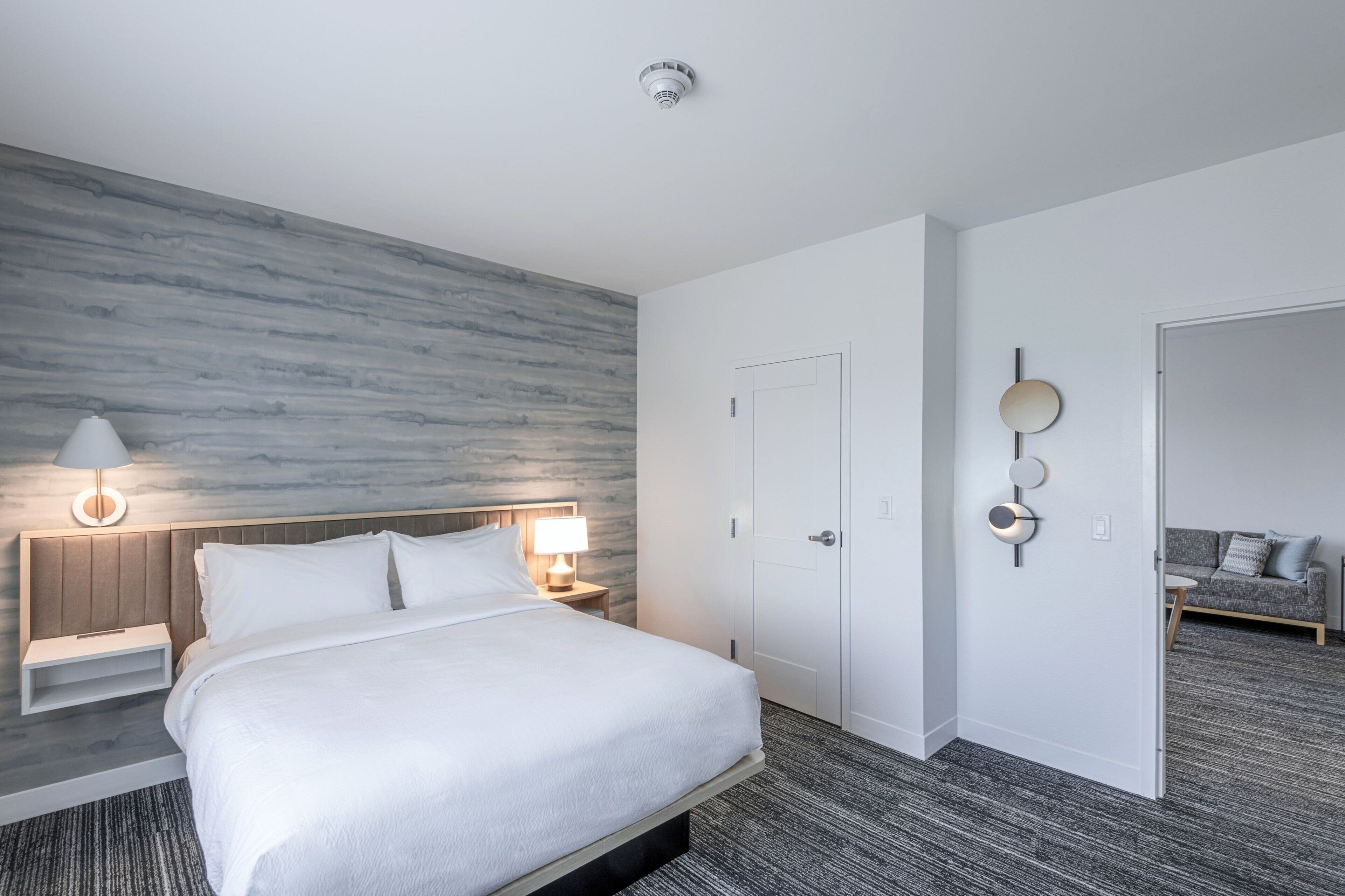 Et moderne hotelværelse med en stor seng, et natbord med lampe, en vægmonteret lysarmatur og en dør, der fører til et andet værelse.