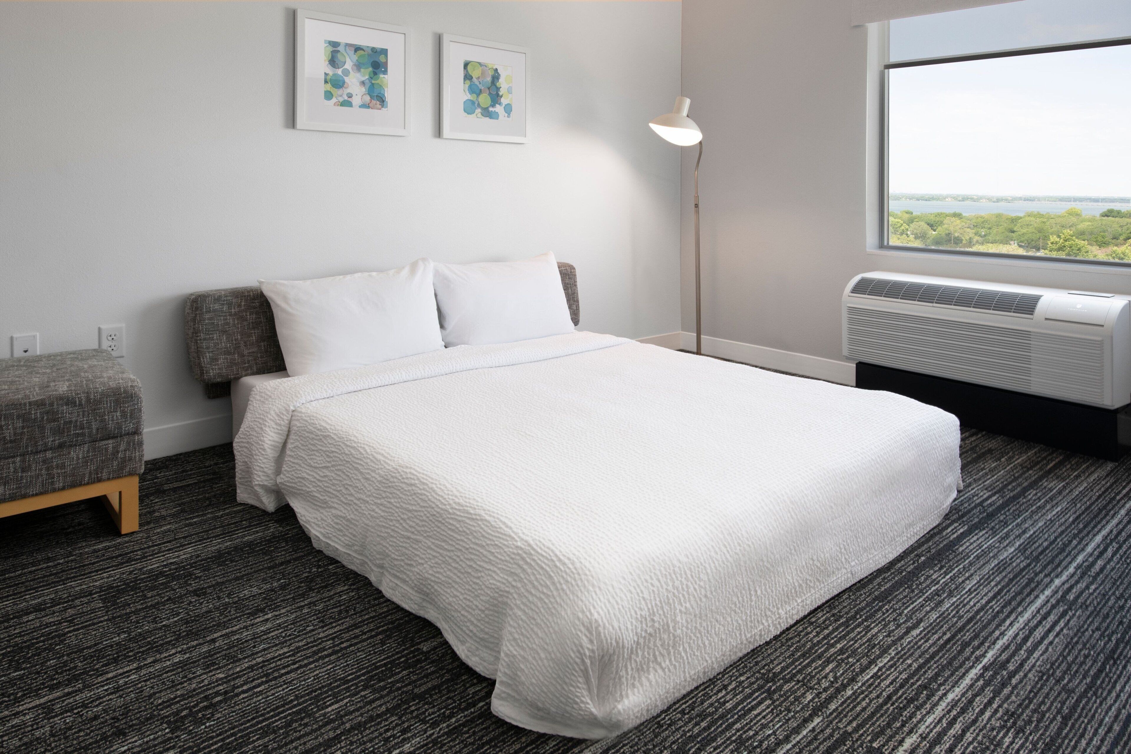 A hotel room with a large bed, a gray armchair, a standing floor lamp, and a window with a view of trees and water.