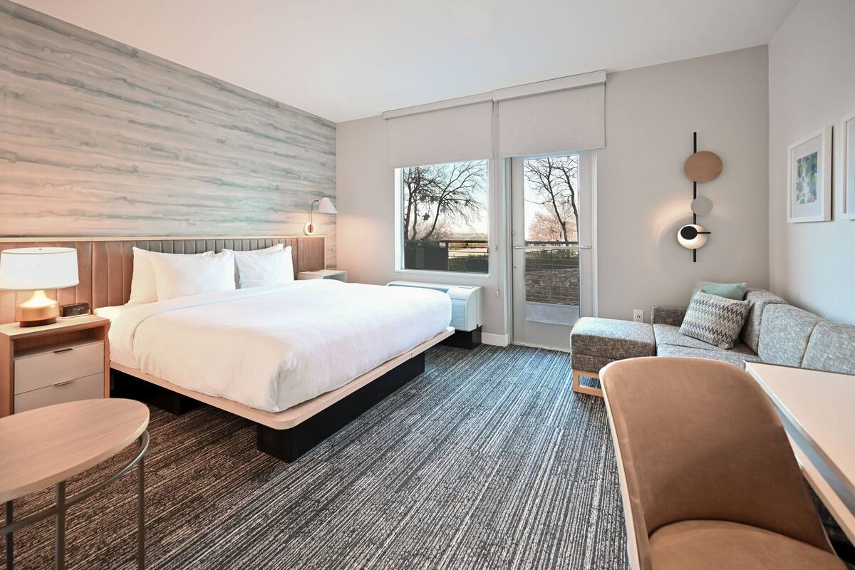 A modern hotel room with a large bed, a sofa, a dining table, and a view of the outdoors.