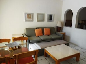 Ein Wohnzimmer mit einer Couch, zwei Sesseln, einem Couchtisch und einer Lampe. An der Wand hängen drei gerahmte Bilder.