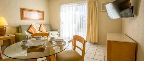 Une chambre d’hôtel avec une table à manger en verre, des chaises en bois, un canapé avec des coussins, un téléviseur à écran plat fixé au mur et une fenêtre avec des rideaux.