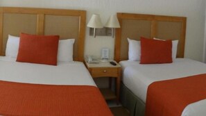Ein Hotelzimmer mit zwei Betten, jeweils mit einem hölzernen Kopfteil, weißer Bettwäsche und roten Kissen. Zwischen den Betten befindet sich ein Nachttisch mit einer Lampe, einem Telefon und einem kleinen Tisch.