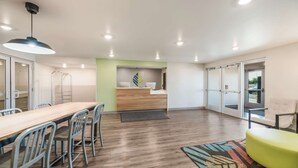 A modern hotel room with a wooden dining table, metal chairs, a green accent wall, a bed with a striped pillow, and a large window. - Woodspring Suites Tucson South (Tucson)
