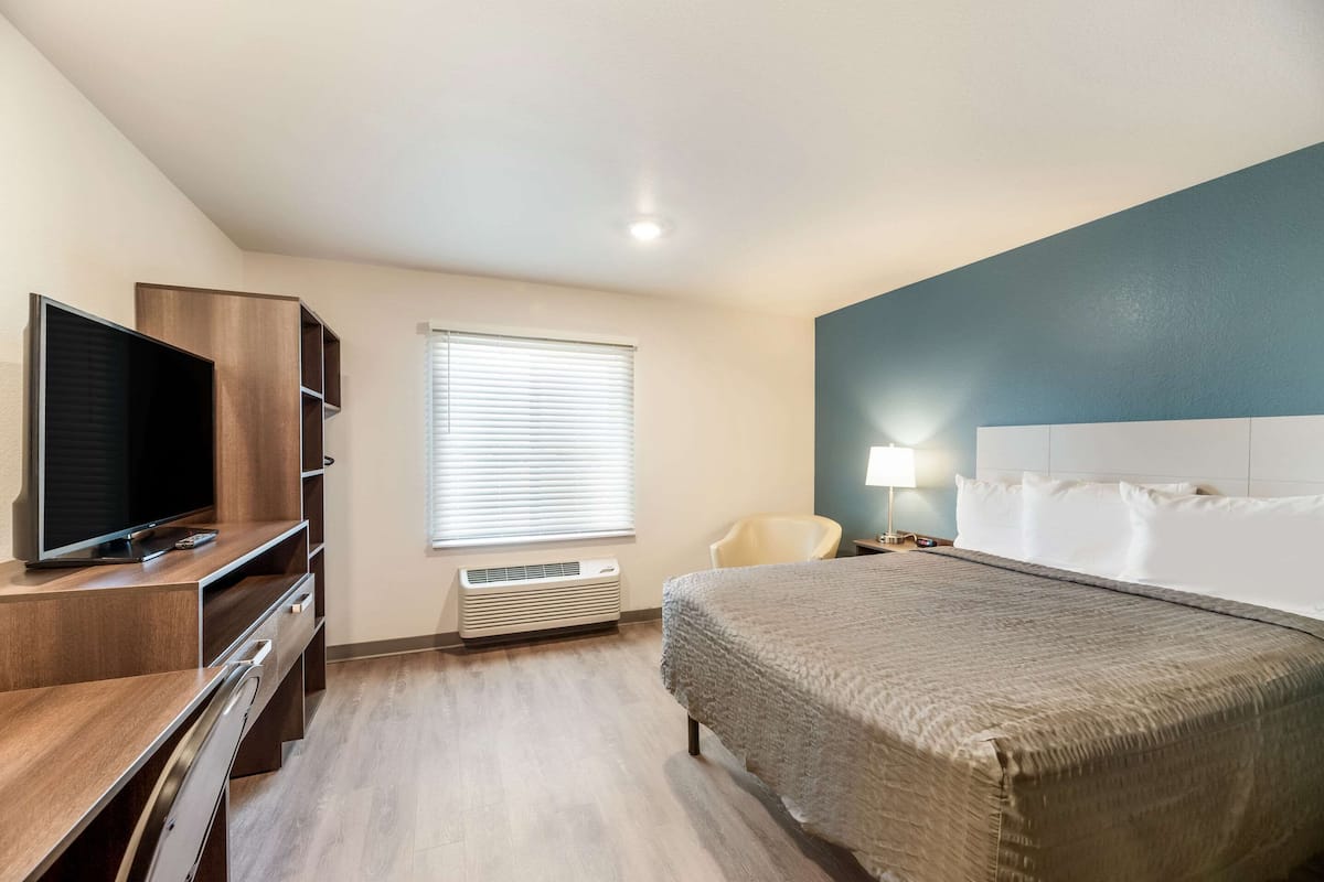 Habitación de hotel con cama, escritorio con televisor, una silla, ventana con persianas y una lámpara fijada a la pared.