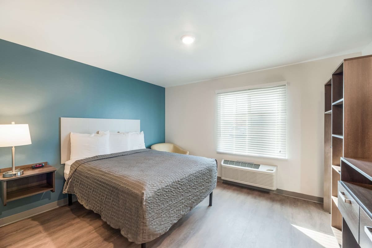 Un dormitorio con cama, mesita de noche, lámpara, equipo de aire acondicionado y ventana con persianas.