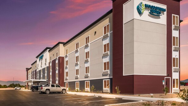 A multi-story hotel with a maroon and beige facade, a prominent logo, and a parking lot with several vehicles. - Woodspring Suites Tucson South (Tucson)