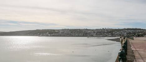 A coastal view with a stone walkway along the water, leading to a town with houses in the background.