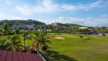 A golf course with palm trees, a red-roofed building, and a hill with buildings in the background.