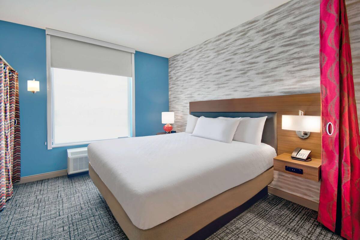 Une chambre d’hôtel avec un grand lit, une table de chevet avec une lampe, un téléphone et une fenêtre avec des rideaux.