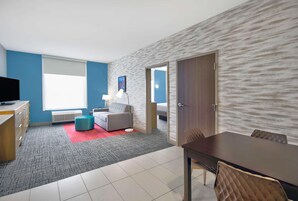 Una habitación de hotel moderna con alfombra gris, sofá gris, mesa de comedor de madera y televisor sobre un aparador de madera.