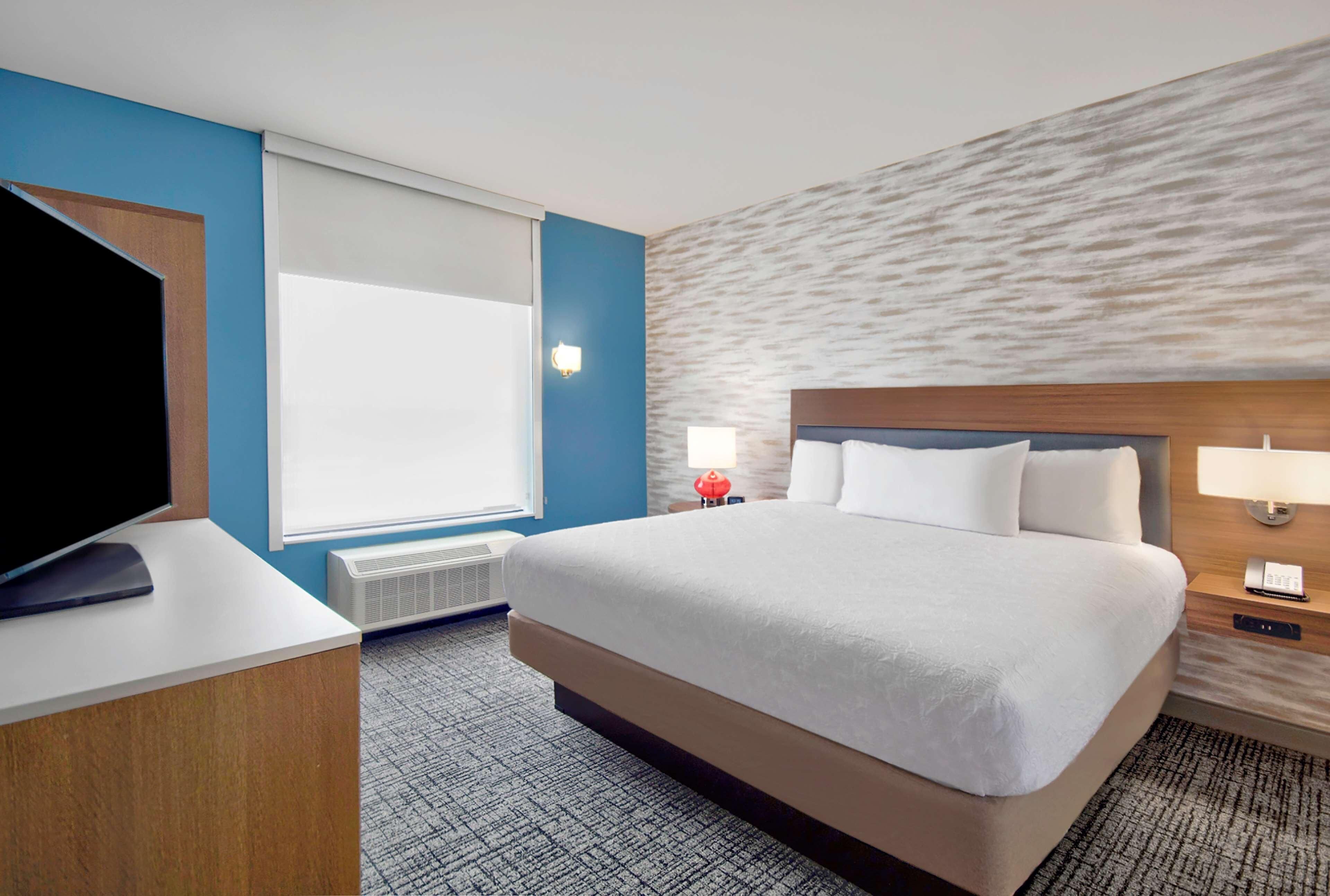 Une chambre d’hôtel dotée d’un grand lit, d’un téléviseur à écran plat, d’une fenêtre avec stores, d’une lampe fixée au mur et d’un mur à texture.